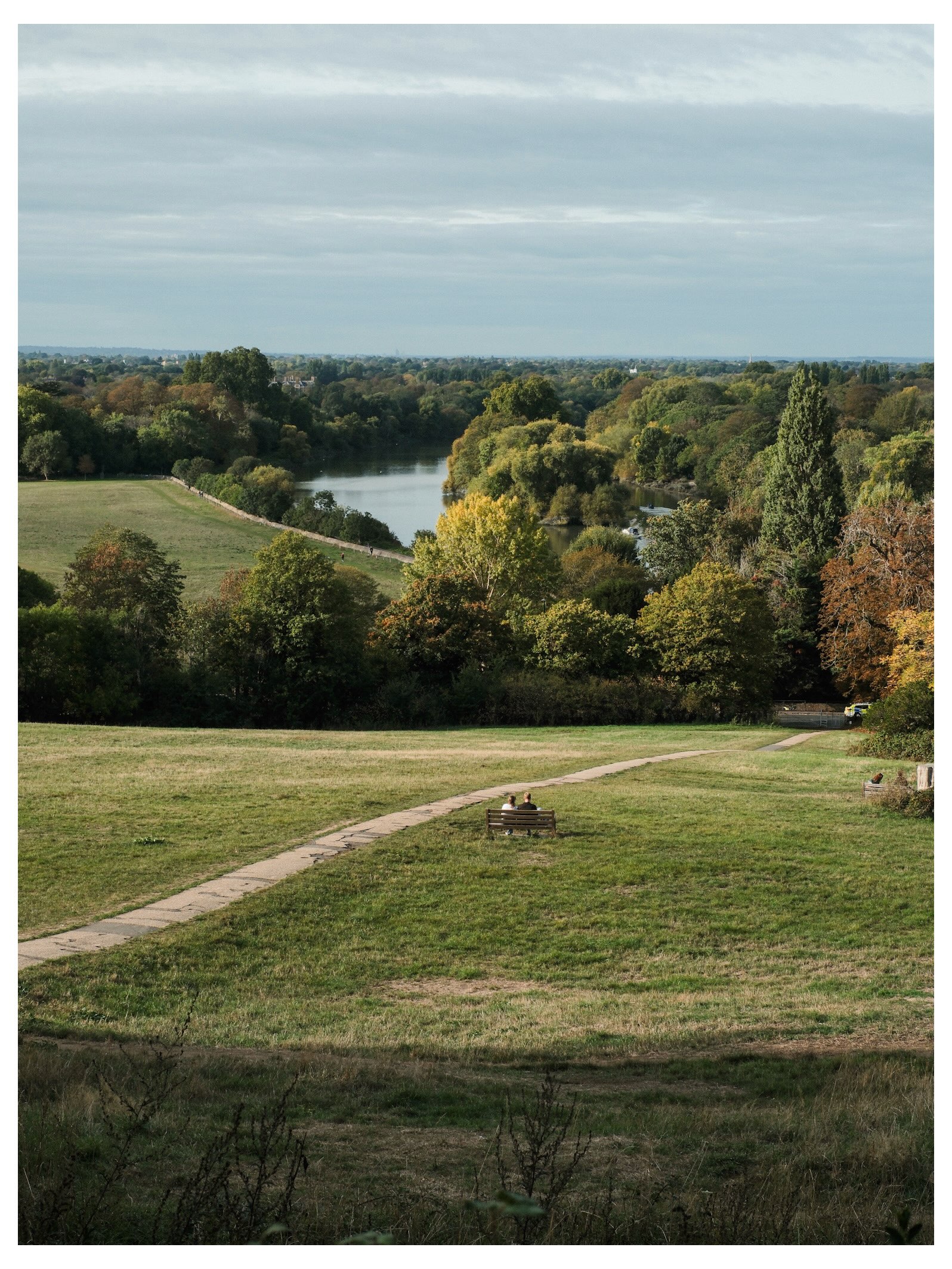 A path through a green park overlooking a lake and autumn trees